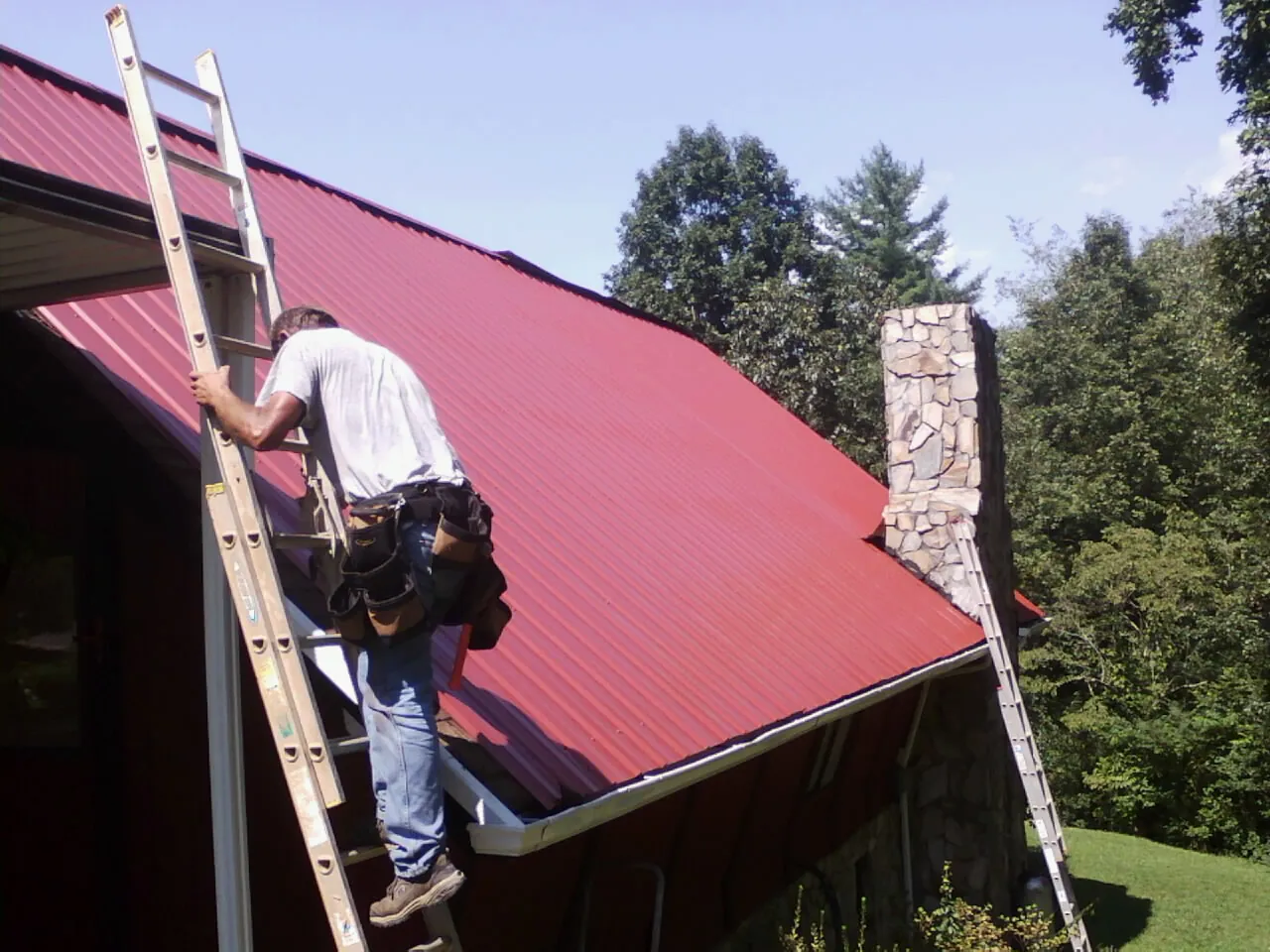 Roofer on a ladder beside a red metal roof and stone chimney — Soffit & Fascia Repair by Roofing Brothers in West Union